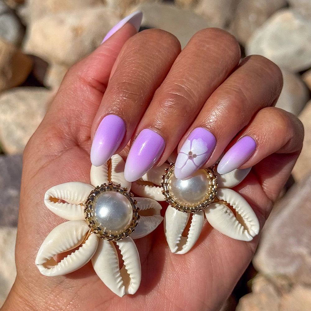A hand with lavender ombré almond nails—styled in glamnetic’s Lilac Reef Press-On Nail Set and detailed with a white flower—holds two cowrie shell and pearl flower earrings against pale rocks. - Product image