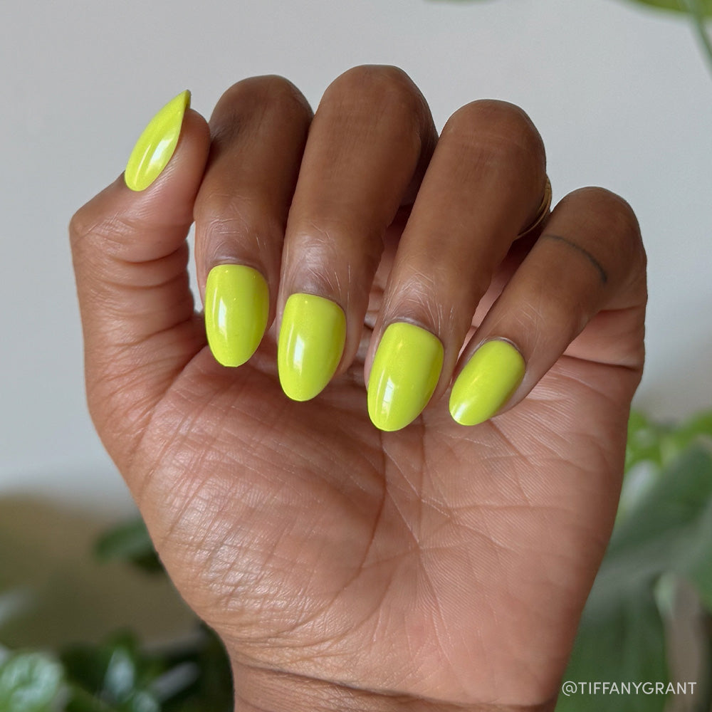 A hand with medium brown skin displays the glamnetic Lime Green short oval nails in a bold neon yellow-green shade, set against a blurred green foliage background. - Product image