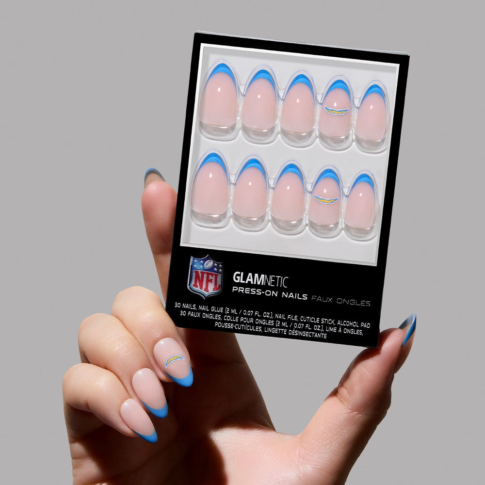A hand holds the glamnetic Los Angeles Chargers Press-On Nail Set, featuring light pink, almond-shaped nails with blue French tips and small Chargers logos—perfect for superfans.
