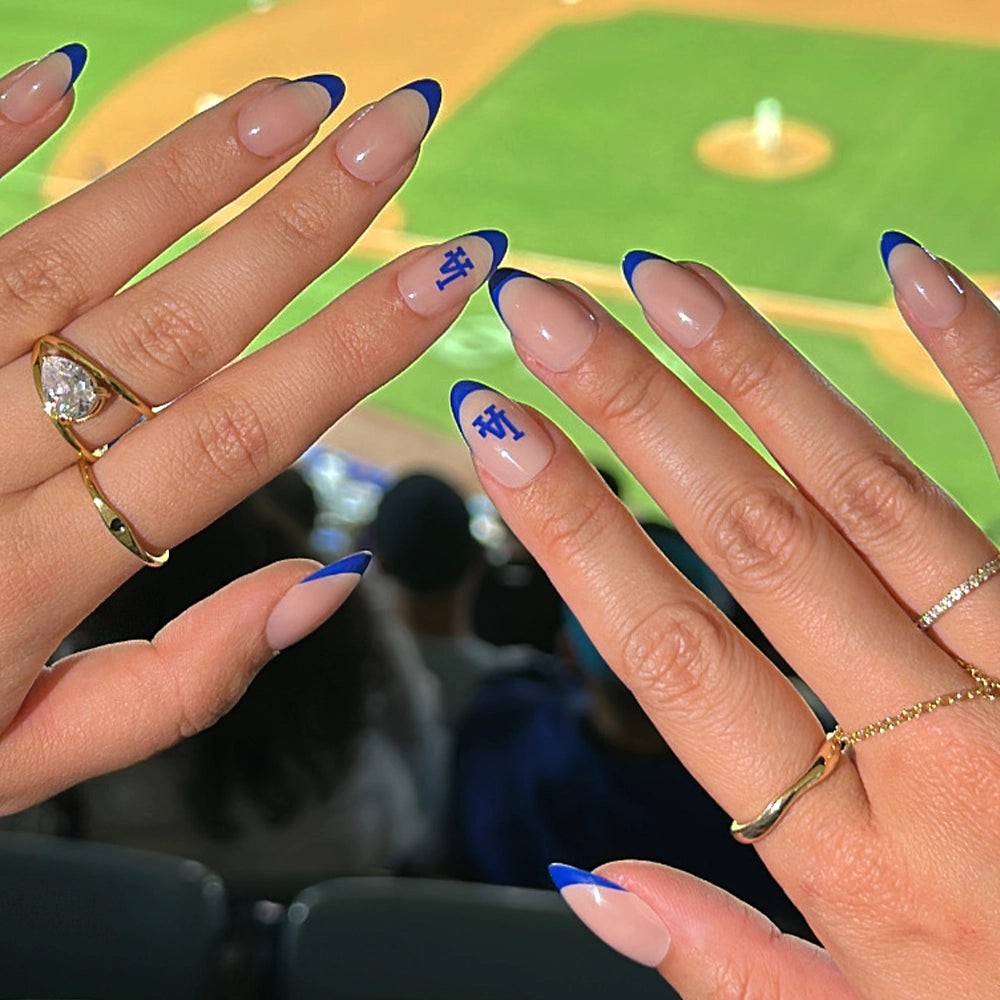 Hands with blue French tip nails and tiny Dodgers characters from the glamnetic Los Angeles Dodgers Press-On Nail Set, accessorized with rings, are shown against a blurred baseball field background.