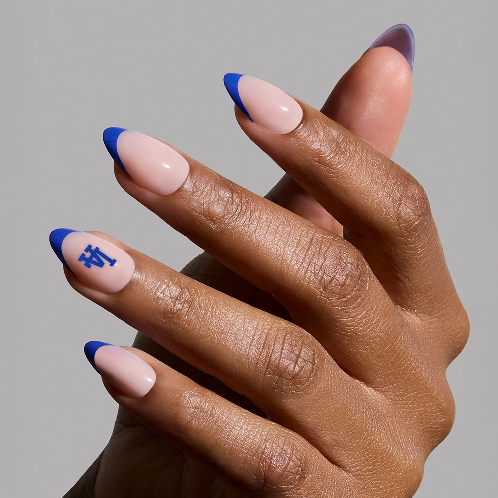 A hand wears the glamnetic Los Angeles Dodgers Press-On Nail Set with a nude base, blue French tips, and a blue LA logo on the ring finger against a plain gray background. - Product image