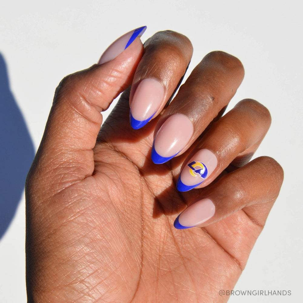 Show off your team spirit with the glamnetic Los Angeles Rams Press-On Nail Set: almond-shaped nails with a nude base, blue French tips, and a gold and blue Rams logo accent. Perfect for fans! Displayed on a white background.