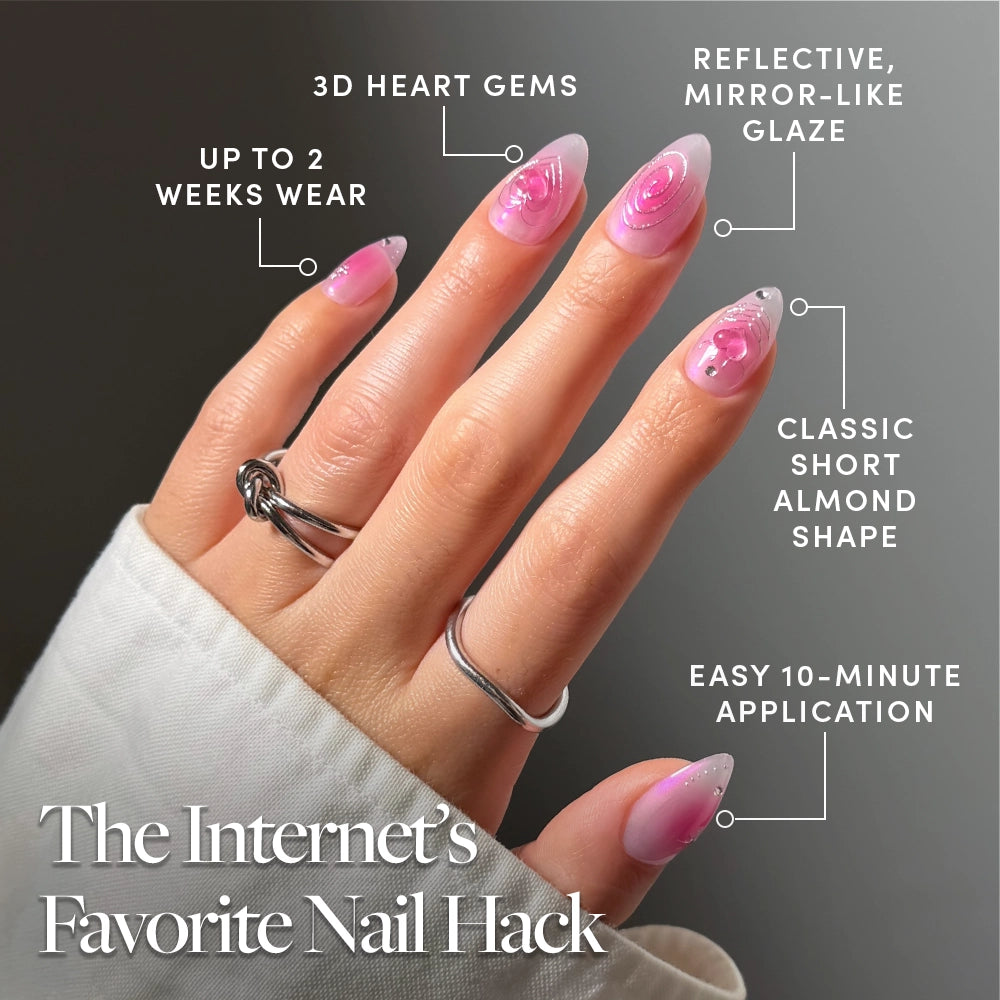 A hand with pink aura “Love Current” nails by glamnetic, shaped into short almonds and accented with 3D heart gems and a reflective glaze. Text highlights their trendy press-on design, up to 2 weeks of wear, and easy 10-minute application. - Product image