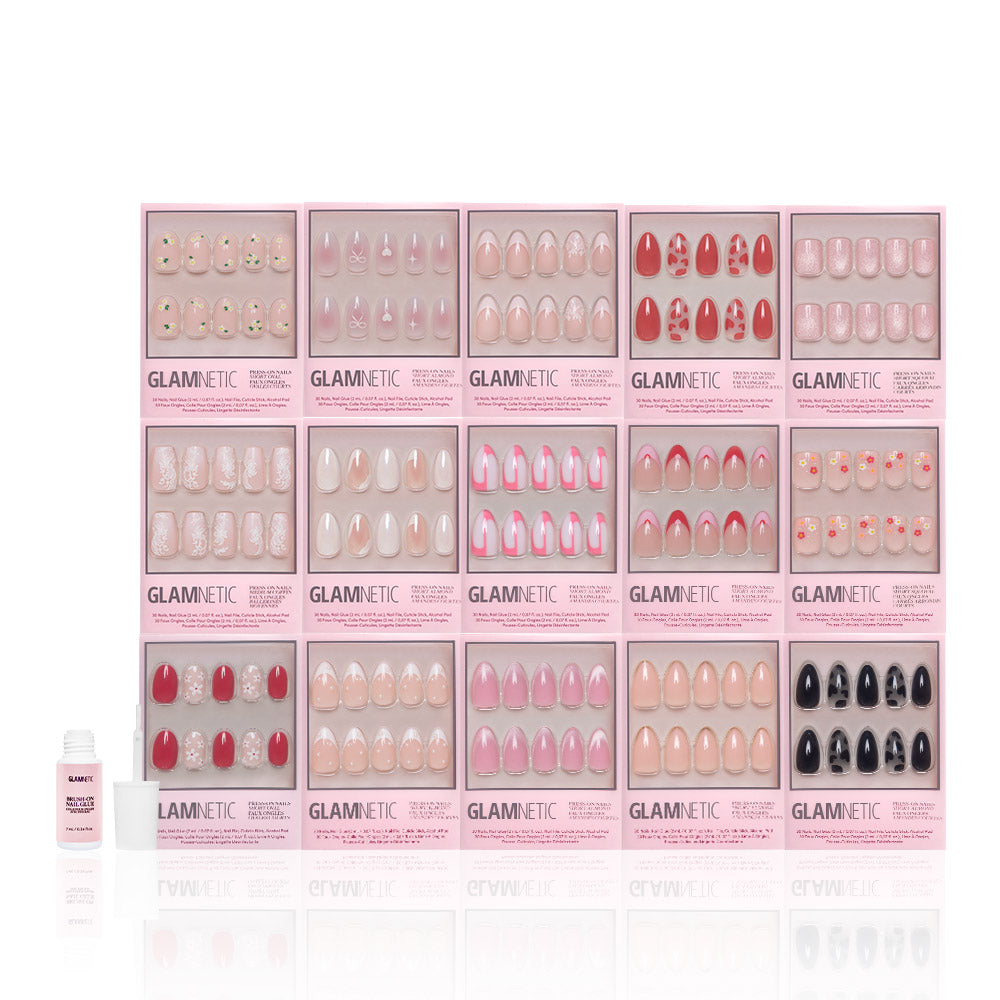 A display of 18 Glamnetic Love In Bloom Collection press-on nail sets, featuring short almond shapes and heart accents in various designs, arranged in three rows with a small bottle of nail glue in front on a white background. - Product image