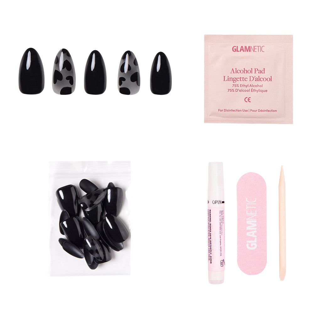 The Love Me Not almond black nails by glamnetic feature cow-print and heart accents, and come with plain black nails, alcohol pad, nail glue, a pink nail file, and a wooden cuticle stick, all arranged on a white background. - Product image