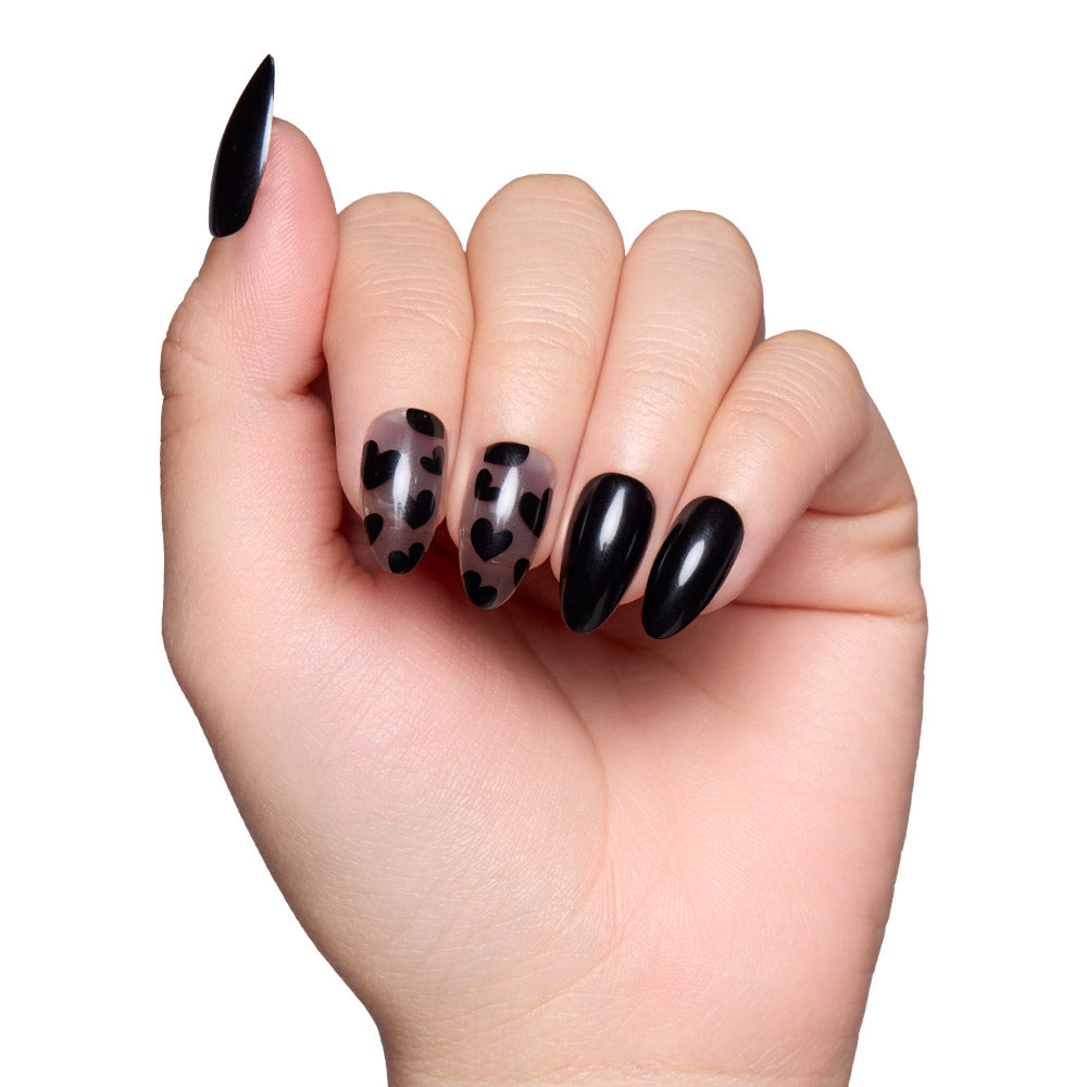 A hand with long glamnetic Love Me Not almond nails is shown; two nails have clear bases with black hearts for an edgy romantic touch, while the rest are glossy solid black. - Product image