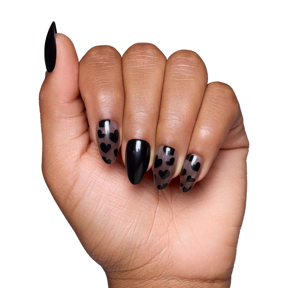 A hand with medium brown skin shows almond manicured nails wearing Love Me Not by glamnetic; three nails have sheer bases with black heart accents for an edgy romantic vibe, and two are painted solid black—all set against a white background. - Product image