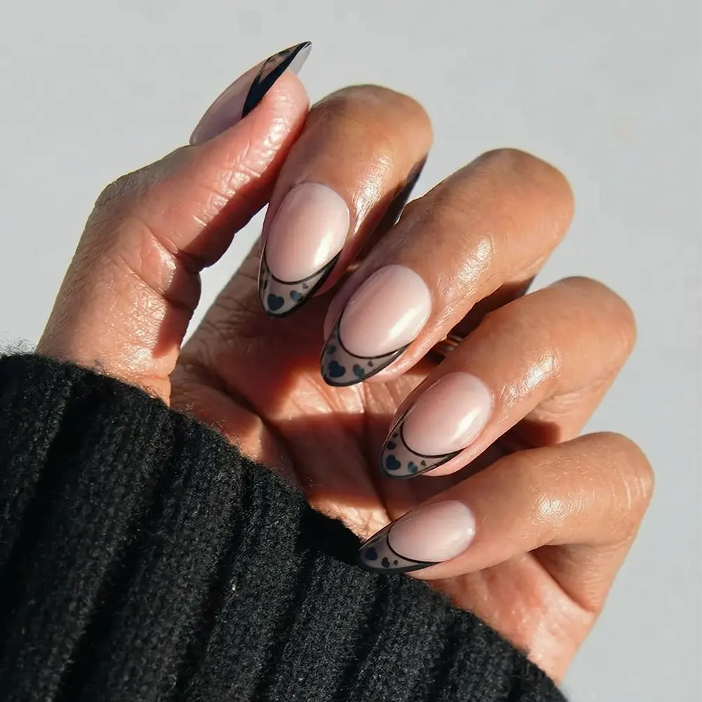 A hand with short almond nails painted in glamnetic’s Love Noir—a nude shade with black abstract French tips and spotted details—paired with a black ribbed sweater.