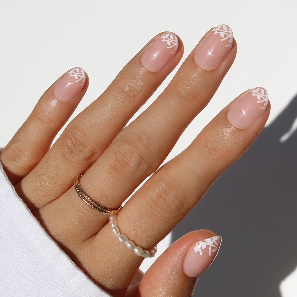 A hand with manicured nails features glamnetic's Love Preserved—natural pink press-ons with white leaf tip designs. The person wears gold and pearl rings and a white long-sleeve top.