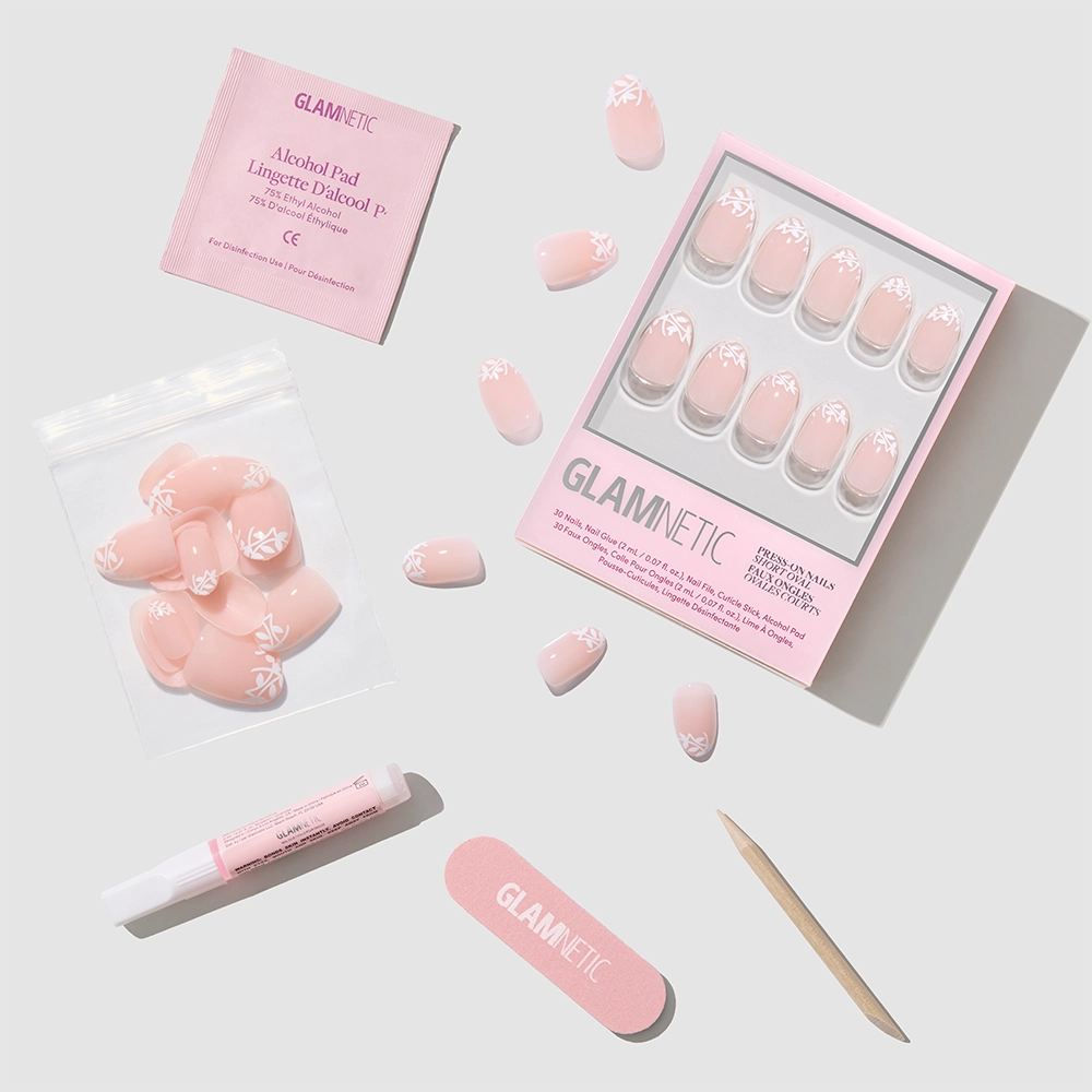 A flat lay of the Glamnetic Love Preserved press-on nail set shows pink botanical nails with a white leaf design, a nail file, cuticle stick, nail glue, alcohol pad, and extra nails in a bag on a light gray surface. - Product image
