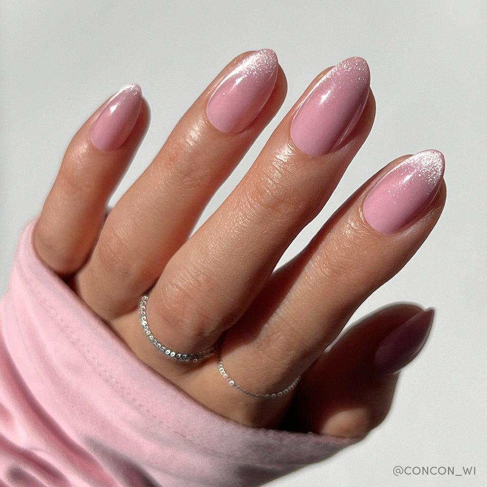 A hand with blush-toned almond nails painted in glamnetic’s Lovers Blush—glossy light pink polish with a subtle white glitter gradient at the tips. The person wears two delicate silver rings and a light pink long sleeve.
