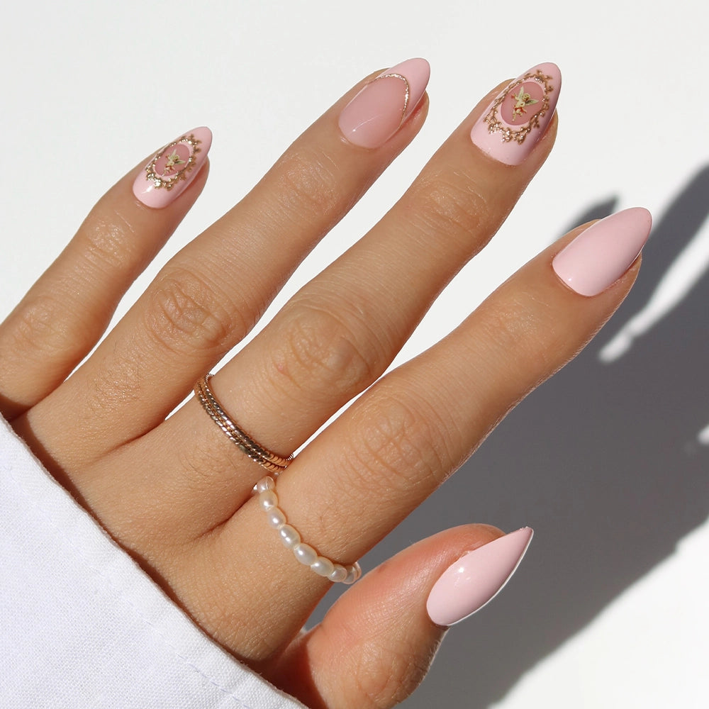 A hand with long, almond-shaped pink nails—Lover's Crest by glamnetic—features Victorian-inspired gold and floral accents on two nails. Three rings are worn: a gold band, a pearl ring, and a twisted gold ring. A white sleeve is visible.