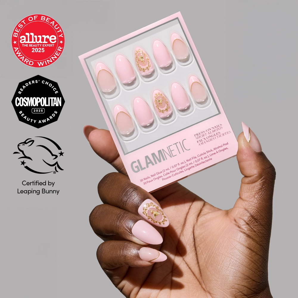 A hand with decorated pink Lover's Crest press-on nails featuring 3D gold metallic nail art holds a glamnetic box, displayed alongside Allure, Cosmopolitan, and Leaping Bunny logos for beauty awards and cruelty-free certification.