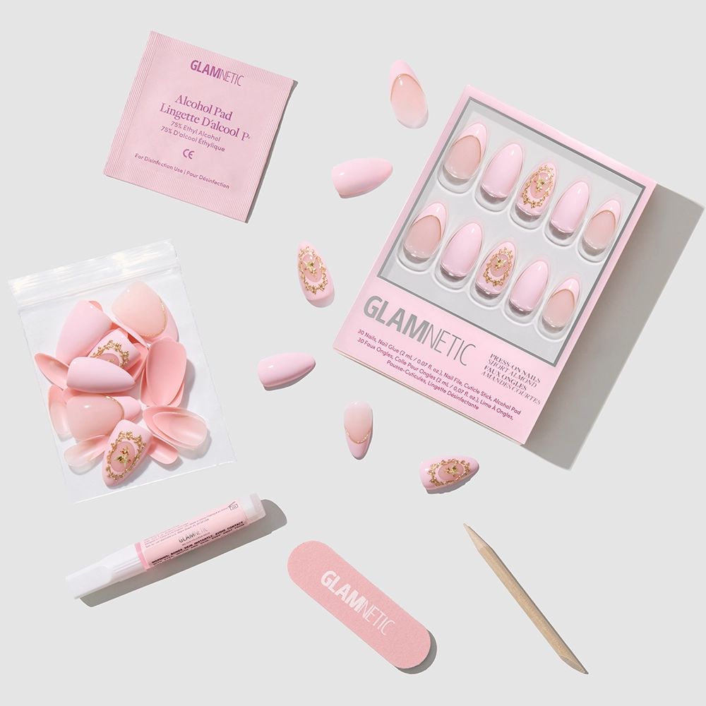 A flat lay of Glamnetic's Lover's Crest press-on nail kit featuring pink nails with Victorian-inspired and 3D gold metallic art, plus a nail file, cuticle stick, glue, alcohol pad, and extra nails arranged on a light background. - Product image