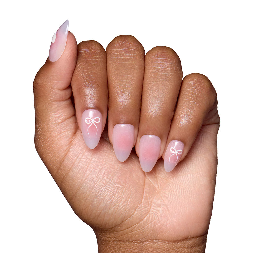 A hand with short almond nails showcases glamnetic’s Lovers Knot: semi-transparent pink ombré nails adorned with delicate raised bows on the ring and middle fingers. - Product image