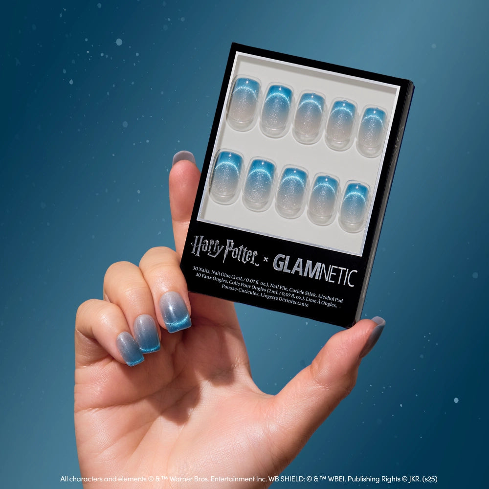 A hand with velvet blue gradient nails from the glamnetic Lumos set holds a box of matching “Harry Potter x glamnetic” nails against a blue background.