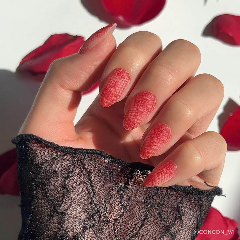 A hand with long, almond-shaped glamnetic Lydia nails featuring a 3D red lace rose design, inspired by Lydia Deetz, rests on a black lace sleeve with scattered red rose petals in the background. - Product image
