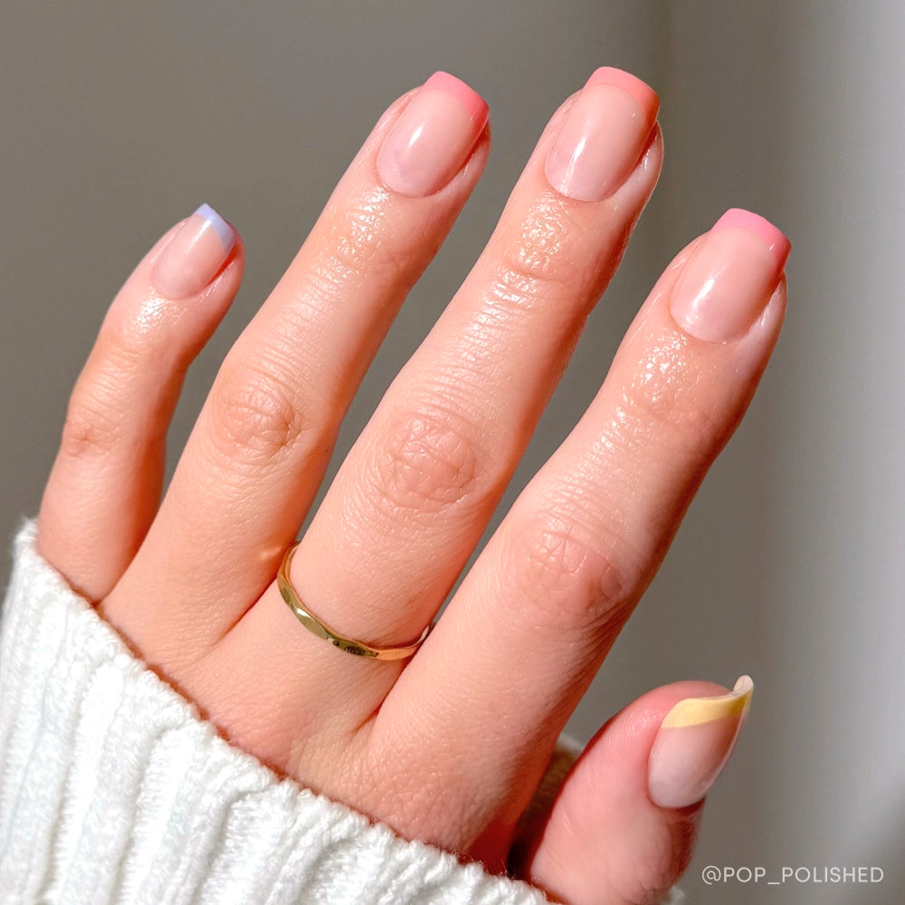 A hand with a gold ring showcases glamnetic Macarons press-on nails, each adorned with a different pastel French tip. The neatly manicured nails pair with a white ribbed sweater against a softly lit, plain background. - Product image