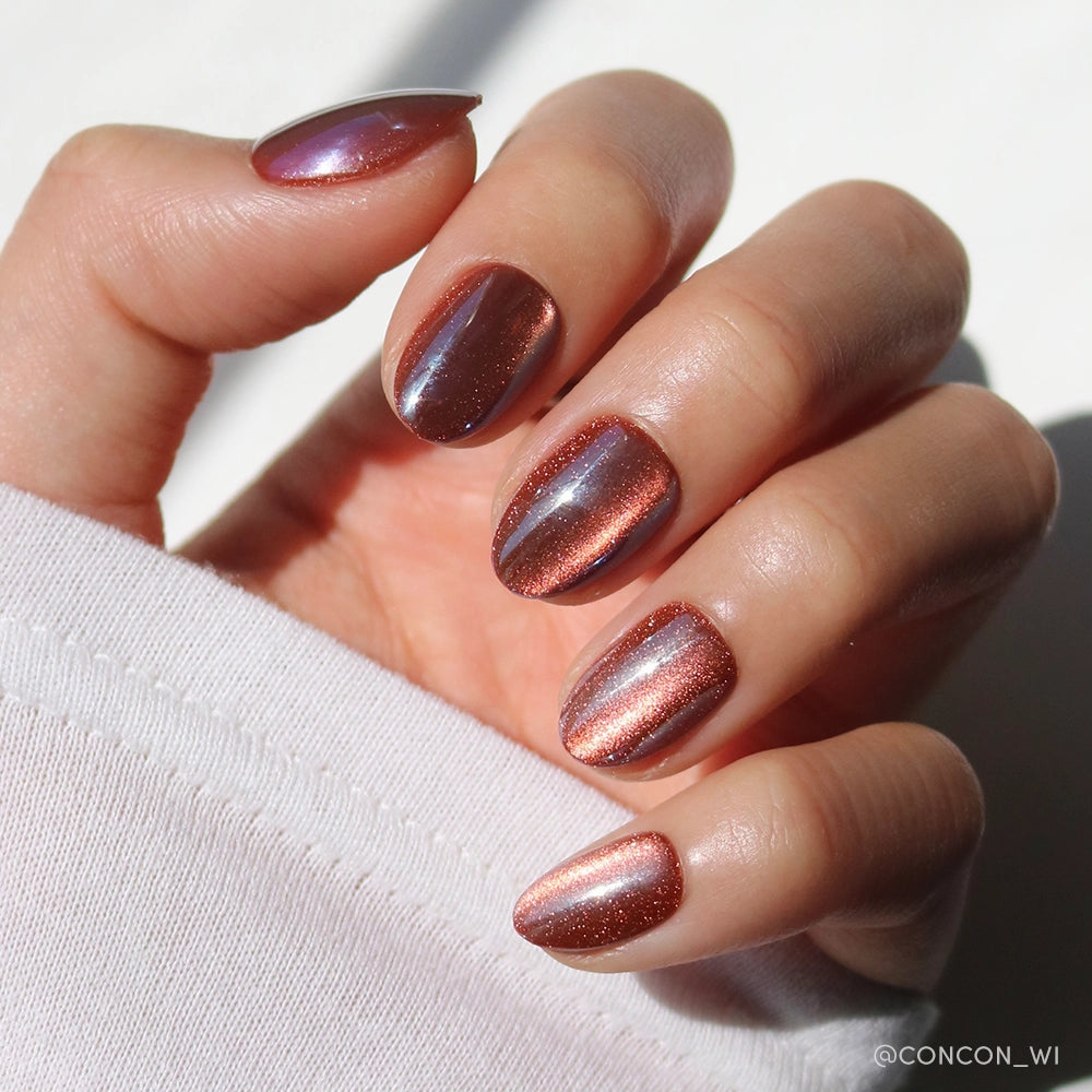 A hand with short oval nails painted in glamnetic’s Magnetic Moscow, a shiny metallic rose gold, glows in sunlight. The person wears a white long-sleeve shirt, highlighting the sets chic elegance.