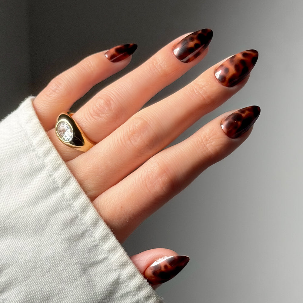 A hand with almond-shaped Main Reference press-on nails by glamnetic in a glossy tortoiseshell pattern, wearing a gold ring with a large oval stone and partly covered by a light-colored long-sleeved shirt. - Product image