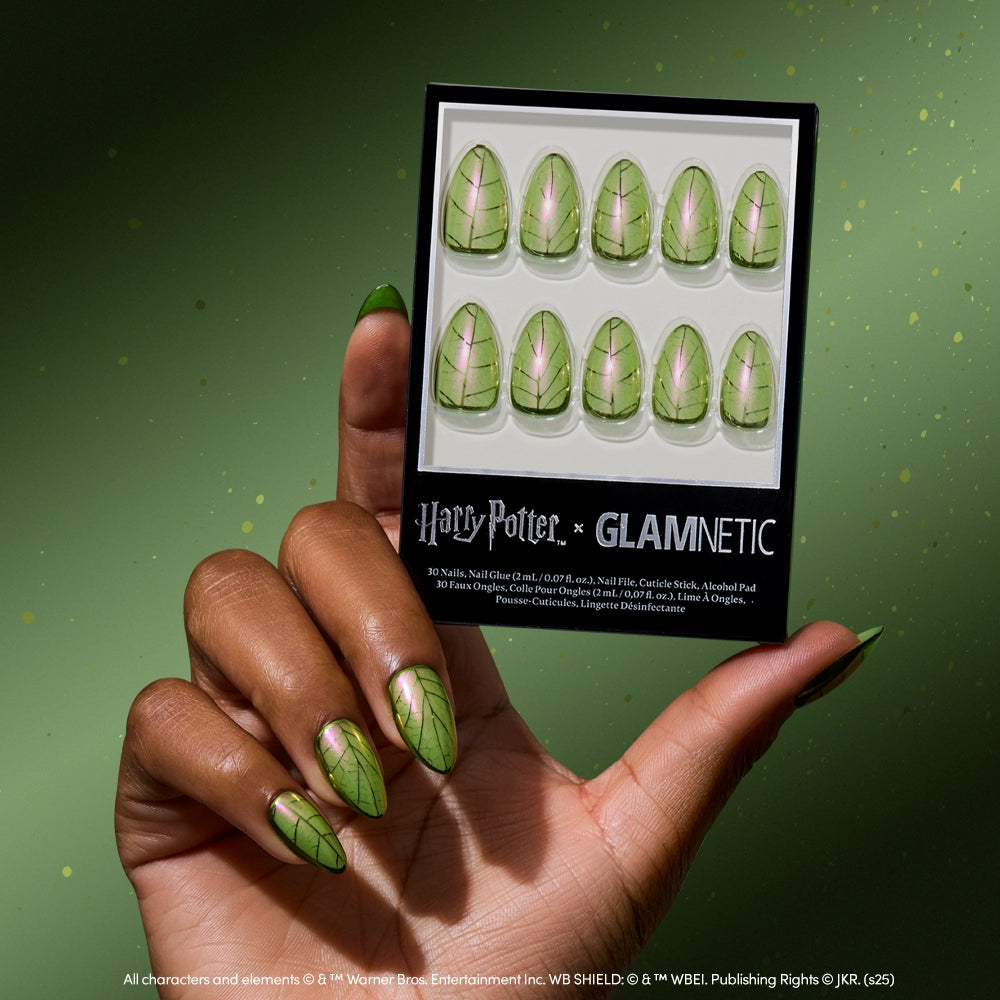 A hand with manicured nails featuring magical green leaf designs holds a box of the limited edition glamnetic Mandrake nail set, decorated with matching artwork, against a green background.