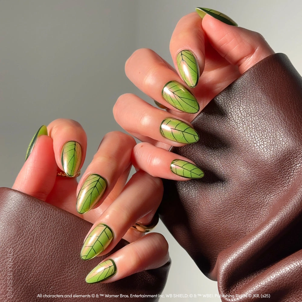 A close-up of hands with green, almond-shaped glamnetic Mandrake nails featuring black leaf vein designs, inspired by magical plants, as the wearer holds their hands together in a brown leather-look jacket. - Product image
