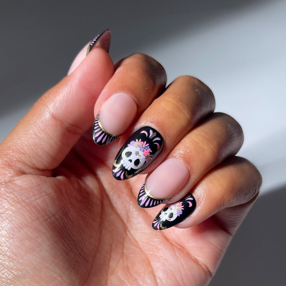 A hand showcases glamnetic’s Mani De Los Muertos press-on nail set, featuring long, glossy black nails adorned with white skulls, pink flowers, and intricate patterns inspired by Day of the Dead. The nails’ smooth shine adds a dramatic flair, while the softly lit background creates an elegant atmosphere. - Product image