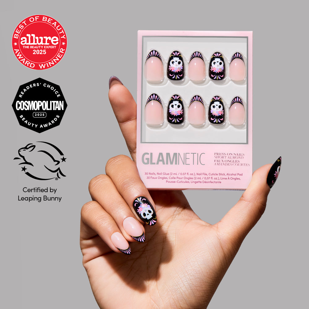 A hand with Halloween-themed, short almond nails holds a box of glamnetic’s Mani De Los Muertos press-on nails. The packaging features a striking skull design inspired by Day of the Dead motifs, along with award logos from Allure and Cosmopolitan, plus a Leaping Bunny certification. - Product image