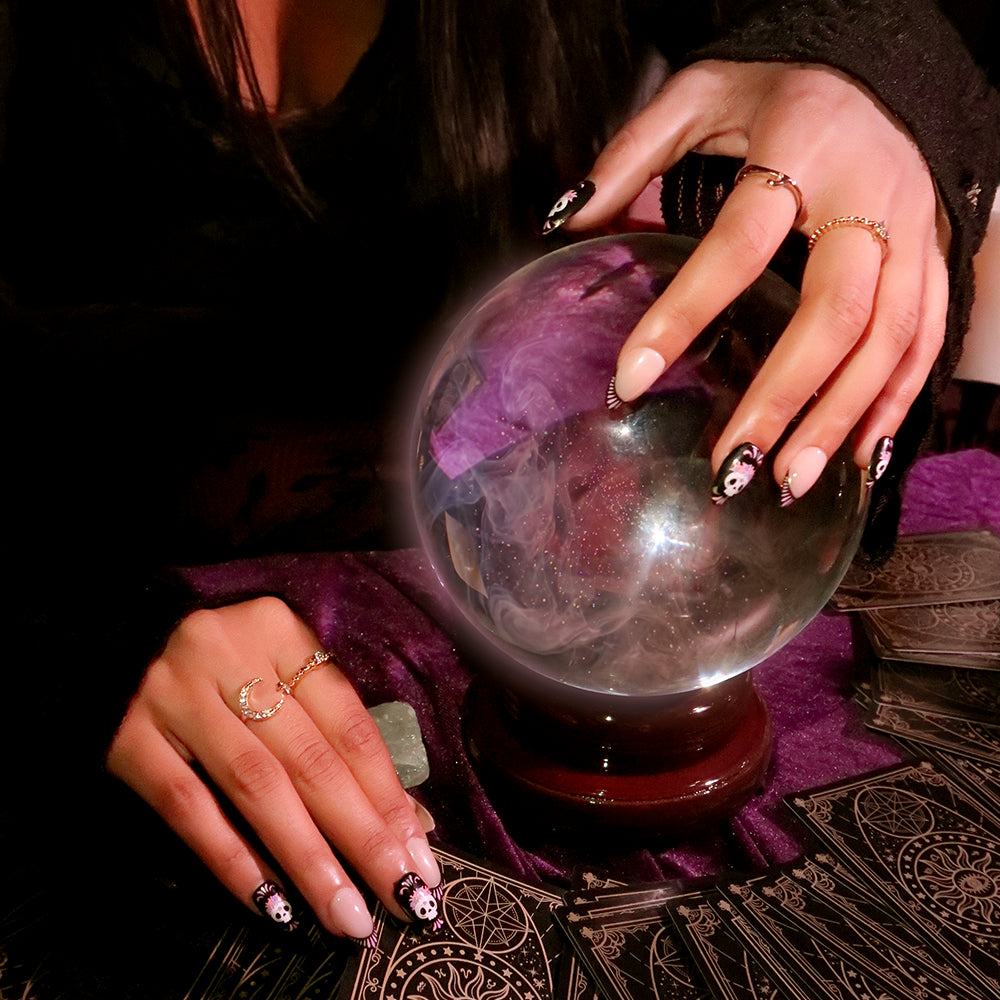 A woman wearing glamnetic’s Mani De Los Muertos nails and rings holds a crystal ball on a purple cloth, surrounded by tarot cards, evoking the atmosphere of a fortune-telling session. - Product image
