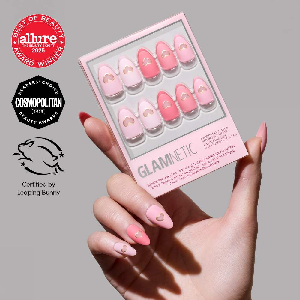 A hand with pink matte "Matte in Love" press-on nails by glamnetic, featuring gold heart cutouts, holds a box of similar short almond nails. The glamnetic box is shown with Allure, Cosmopolitan, and Leaping Bunny awards.