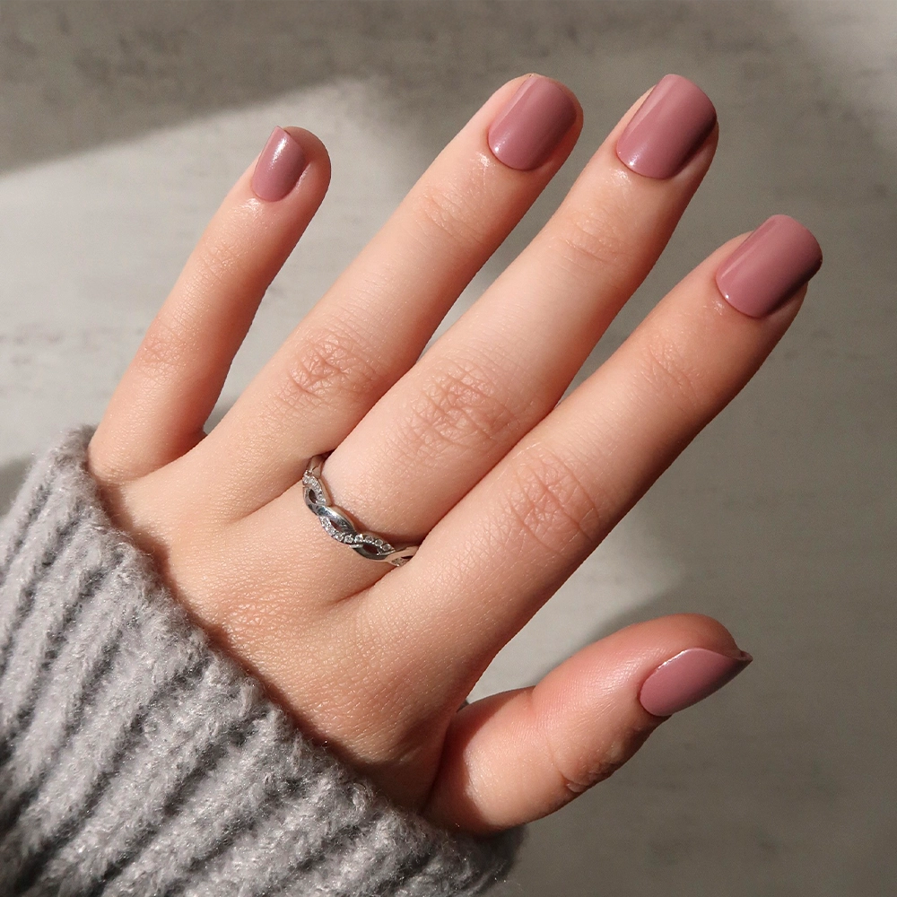A hand with super short, square-shaped nails painted in glamnetic's Mauve Cutie polish, wearing a silver ring with small stones and a gray ribbed sweater, is shown against a light background. - Product image
