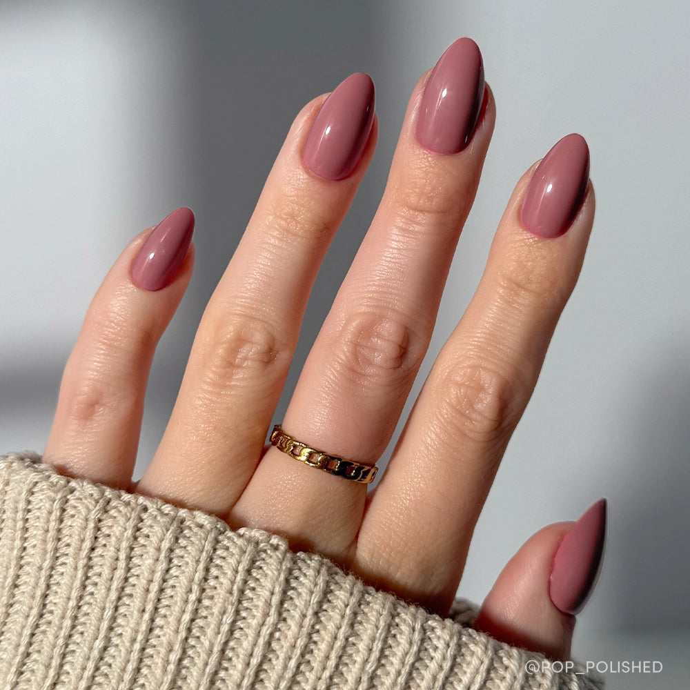 A hand with neatly manicured, short almond nails painted in glamnetic's Mauve Obsession—a dusty rose polish with a glossy finish—wears a gold chain ring and is partially covered by a beige knit sweater sleeve. - Product image
