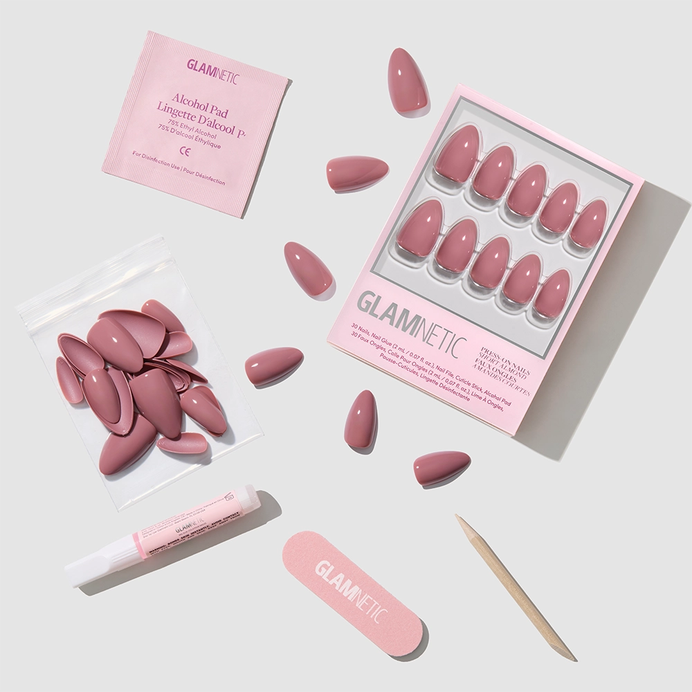 A flat lay of glamnetic's Mauve Obsession press-on nails in a glossy mauve finish, with nail glue, file, wooden cuticle stick, alcohol pad, and extra short almond nails arranged neatly on a light grey background. - Product image