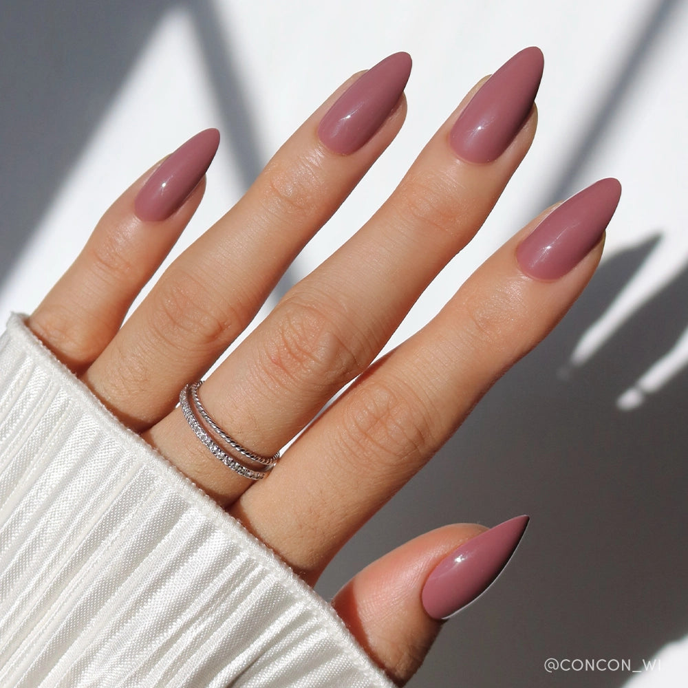 A hand with almond-shaped glamnetic Mauve Wife press-on nails, wearing stacked silver rings, rests on white ribbed fabric. Soft natural light and shadows add a minimal, elegant touch. - Product image