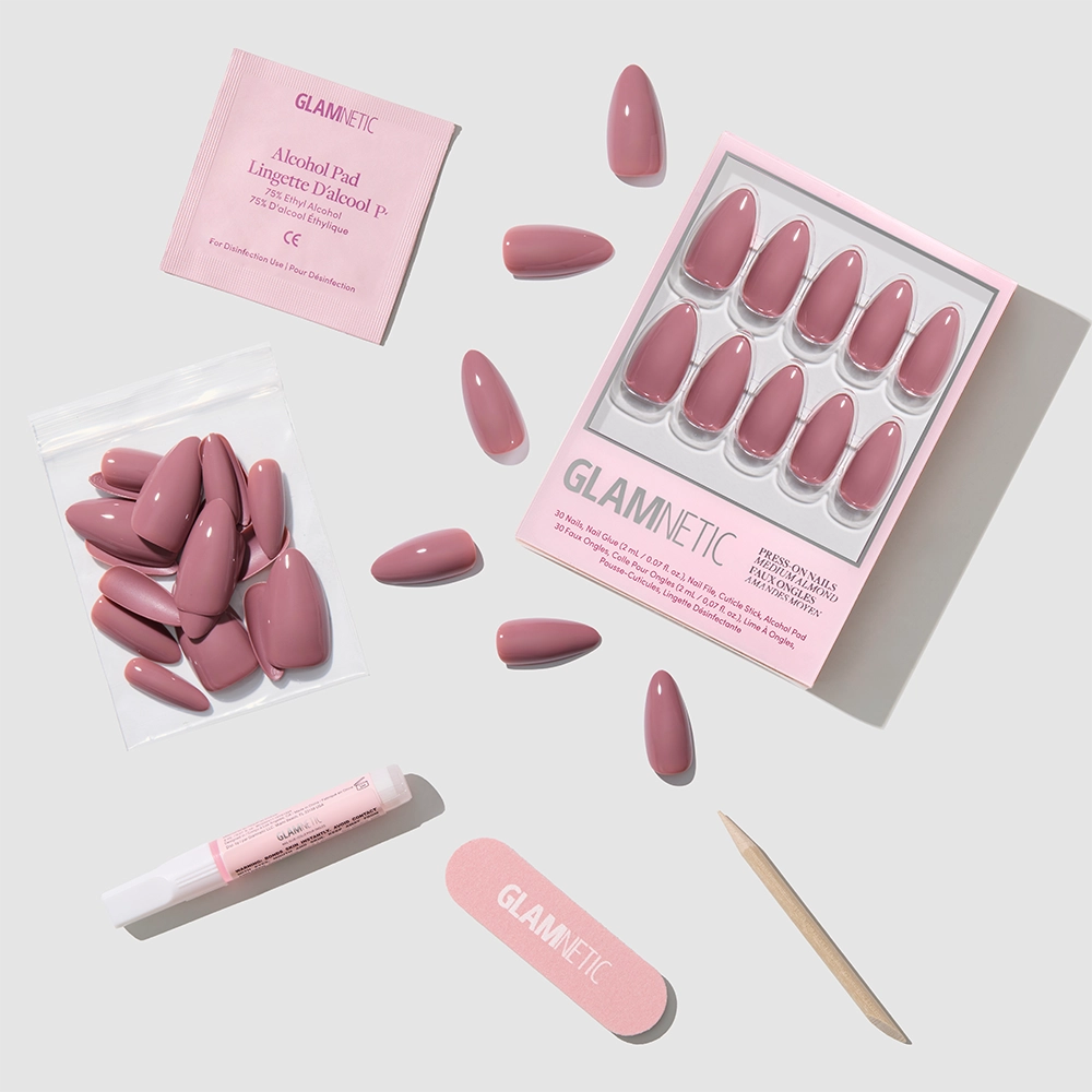 A set of Mauve Wife glossy pink fake nails, adhesive tube, wooden cuticle stick, pink glamnetic nail file, alcohol pad, and extra almond nails are arranged neatly on a light background. The packaging and file show the glamnetic brand. - Product image