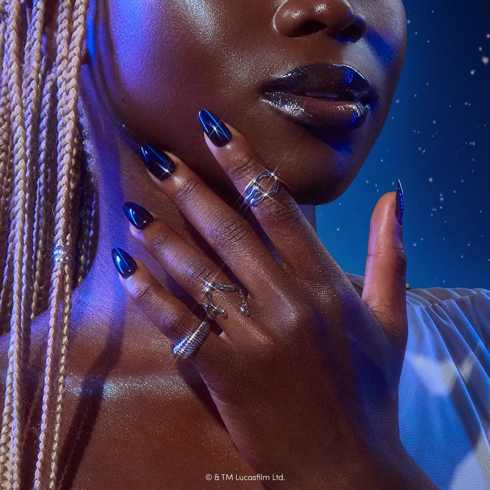 A person with braided blond hair and glossy lips poses with dark nails featuring the glamnetic May The Force Be With You STAR WARS press-on nails design and silver rings on their fingers, set against a starry blue background. - Product image