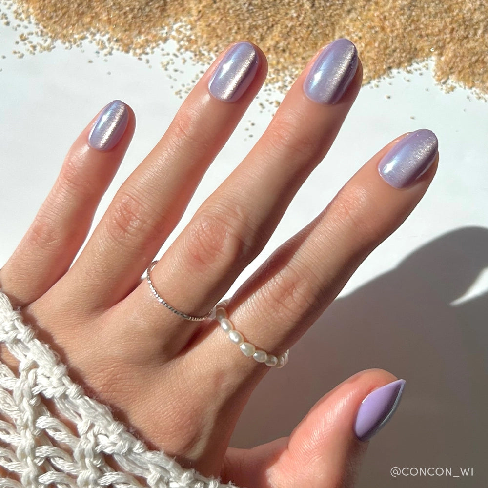 A hand wears glamnetic’s Mermaids Tail iridescent purple press-on nails, paired with silver and pearl rings, resting on white crochet fabric with golden beads scattered in the background. - Product image