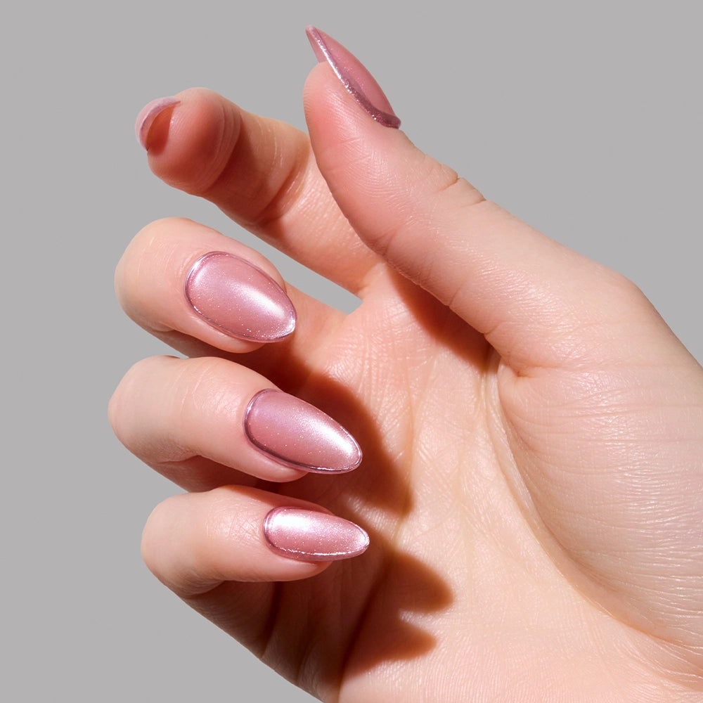 A hand with long, almond-shaped Mertini press-on nails by glamnetic, painted in shiny light pink with subtle foil accents, is held against a plain grey background. - Product image