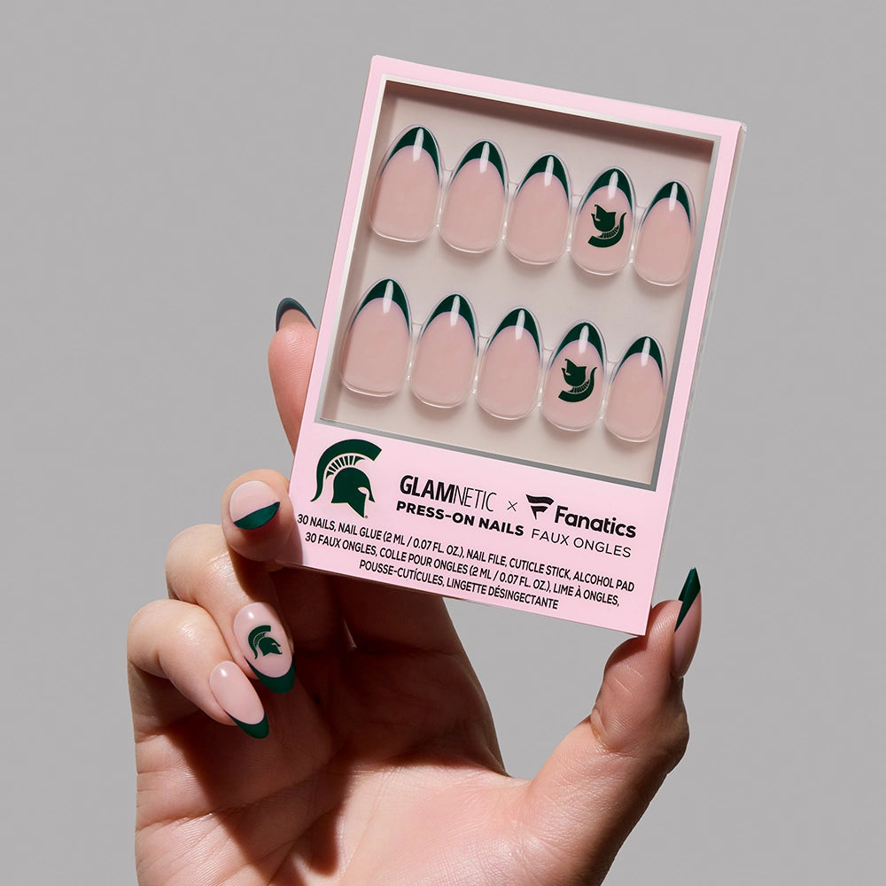 A hand with green-tipped press-on nails holds the glamnetic Michigan State Spartans Press-On Nail Set, featuring the team logo and green French tips on a neutral base.