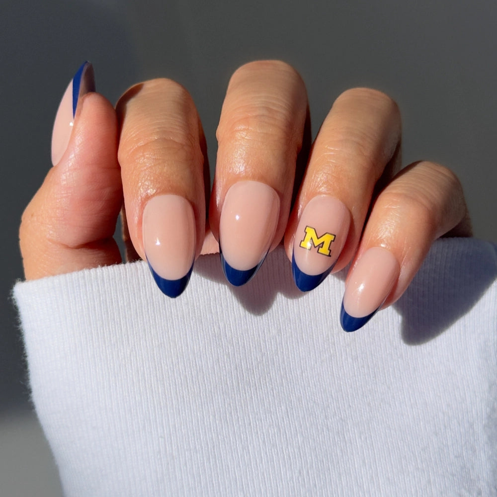 A hand with almond-shaped nude nails, blue French tips, and the Michigan M logo on the ring finger—perfect for flaunting your glamnetic Michigan Wolverines Press-On Nail Set on game day.