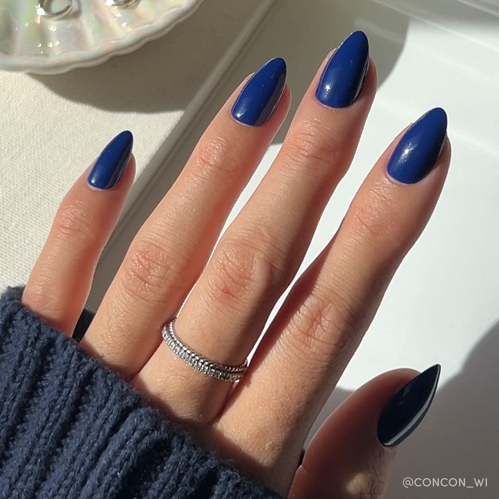 A hand with long, almond-shaped nails in glamnetics glossy, vegan & cruelty-free Midnight Blue set. The person wears a dark sweater and silver double-band ring as sunlight highlights the nails. - Product image