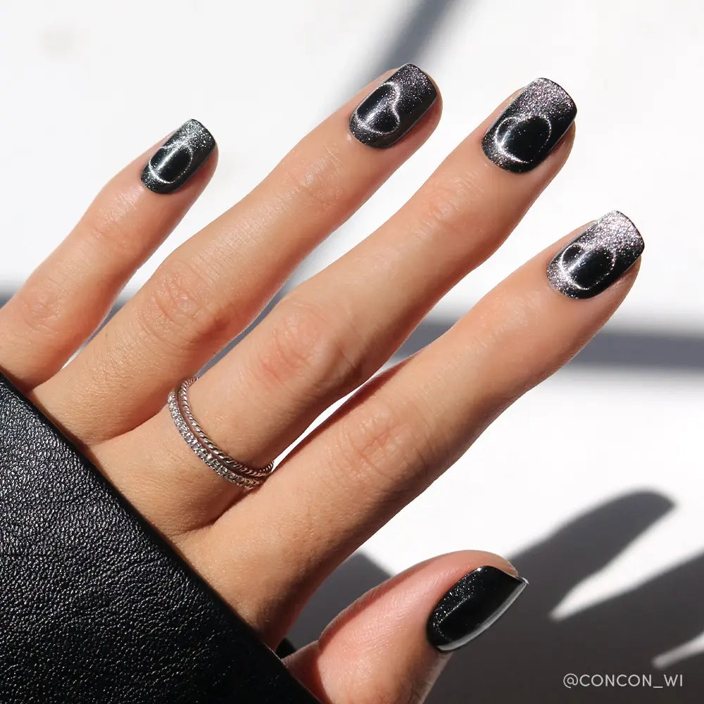 A hand with short, square-shaped nails painted in glamnetic Midnight Romance—a black polish with a silver cat-eye effect—wears a double-banded silver ring, partly covered by a black sleeve. - Product image