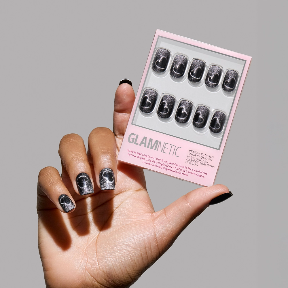 A hand with black and silver crescent moon-themed short squoval press-ons holds a box of glamnetic Midnight Romance nails against a plain gray background.