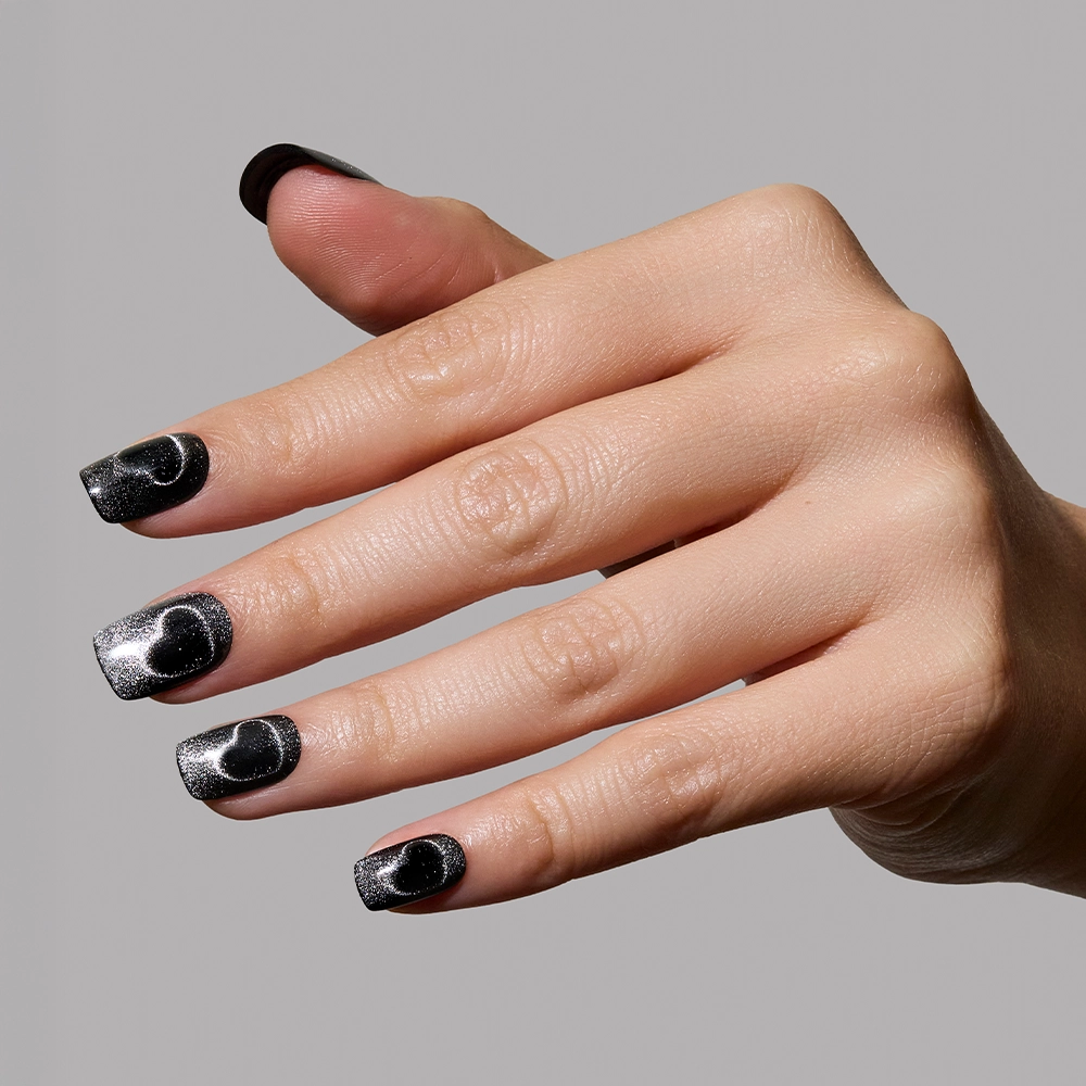 A hand displays glamnetics Midnight Romance: long, square magnetic nails in black with a metallic silver ombre and black heart designs, set against a plain gray background. - Product image