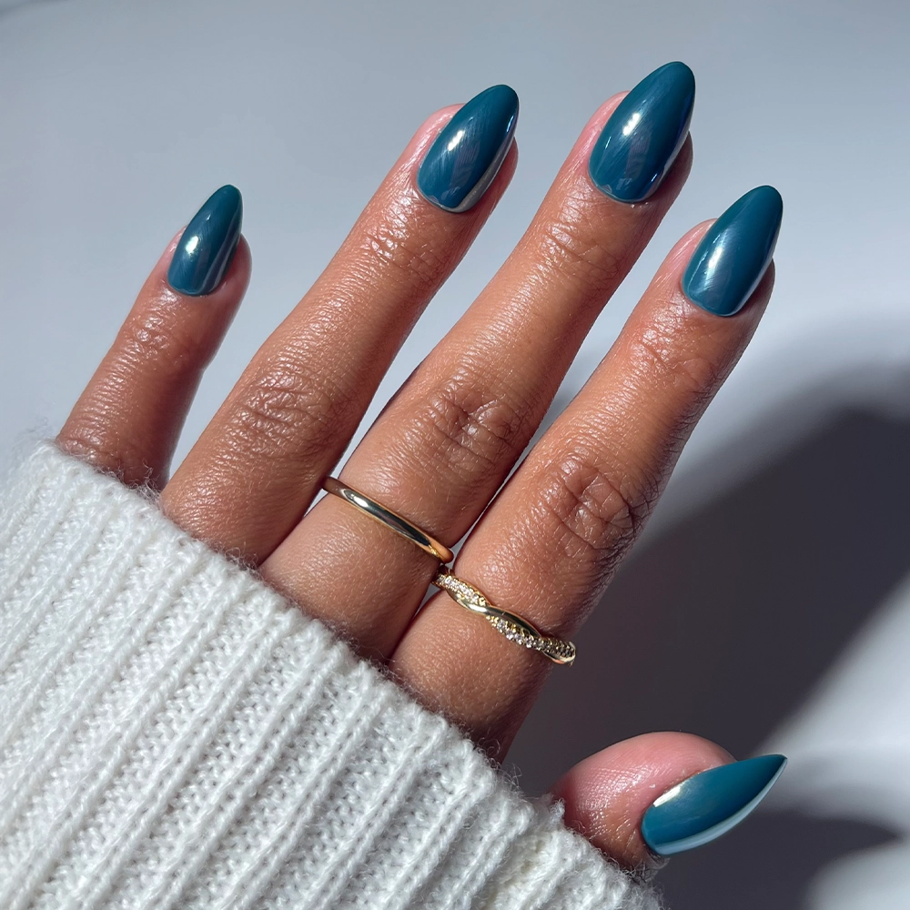 A hand with glamnetic Midnight Teal Press-On Nails in a short almond shape rests on a white surface. The person wears a white ribbed sweater and two gold rings, creating a chic, salon-quality mani look.