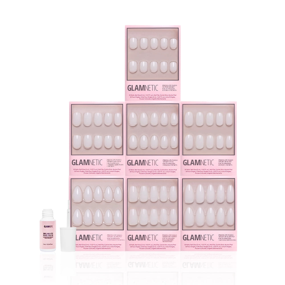 Eight pink boxes of the glamnetic Milky Manis Bundle are stacked in a pyramid, each showcasing neutral milky press-on nails. In front on the left, a small pink bottle of nail glue with an open cap is displayed. - Product image