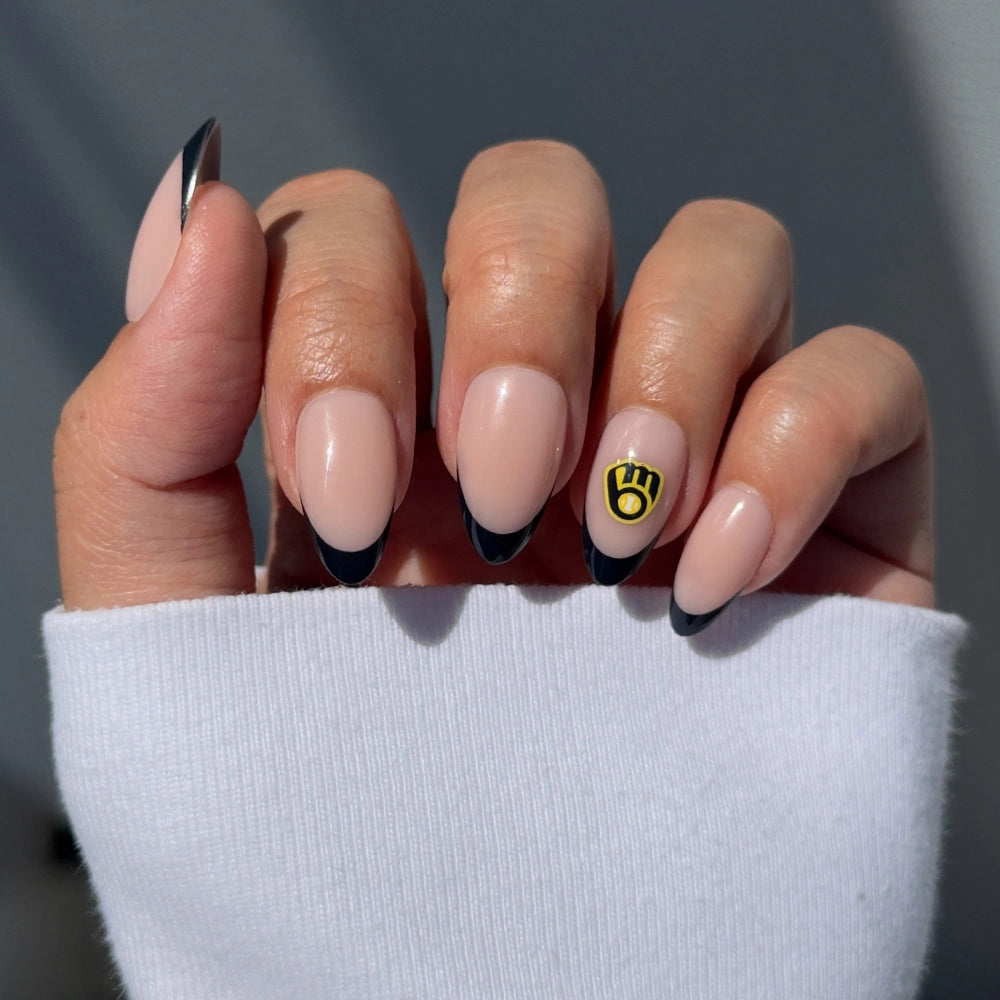 A hand sports the glamnetic Milwaukee Brewers Press-On Nail Set, featuring almond-shaped nude nails with black French tips and a yellow Brewers logo accent on the ring finger. A white sleeve partially covers the hand.