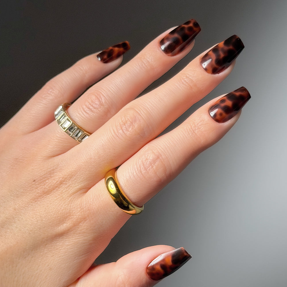A hand displays medium coffin Mine By Right press-on nails by glamnetic in a glossy tortoiseshell pattern, styled with two gold rings—one with rectangular stones, one plain band—set against a neutral background for a salon-quality manicure. - Product image