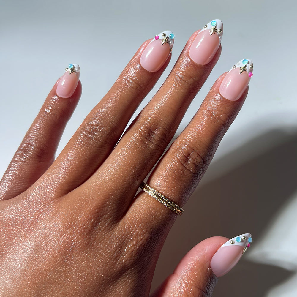 A hand adorned with a gold ring showcases manicured nails featuring a nude base and vibrant, jeweled white floral designs at the tips, reminiscent of the dazzling, jewel-encrusted look of glamnetic's Moonstone press-on nails.
