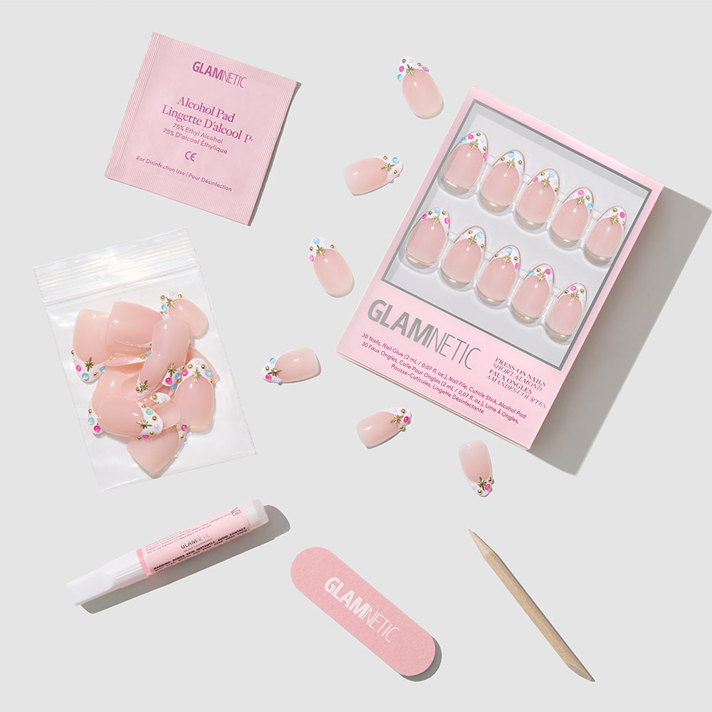 The glamnetic Moonstone press-on nails kit is shown with floral and Moonstone nails, an alcohol pad, nail glue, wooden stick, nail file, and extra nails, displayed against a light gray background. - Product image